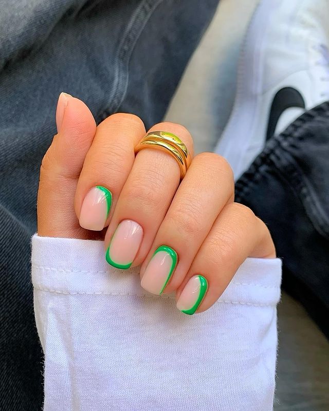 A hand with short, square nails featuring bright green wavy French tips, styled with a white sleeve and a gold ring.