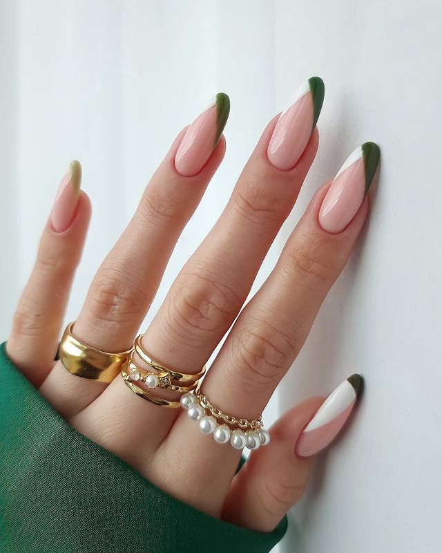 A hand with long, almond-shaped nails featuring dark green and white French tips, accessorized with several gold rings.
