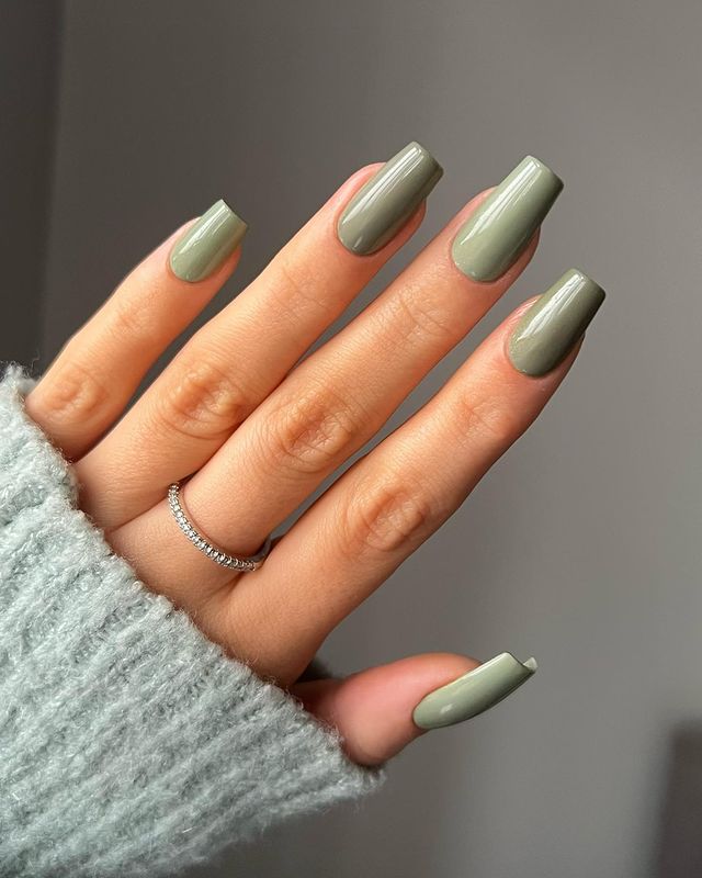 A hand with long, square-shaped nails painted in a glossy olive green color, wearing a light blue knit sweater and a thin diamond band.