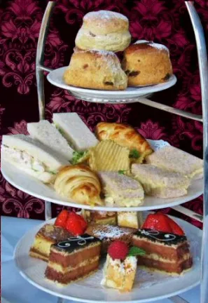 A silver three-tiered serving stand set against a dark red damask background. The top tier holds three golden-brown scones, the middle tier features assorted finger sandwiches and small croissants, and the bottom tier is filled with sliced layered cakes topped with fresh strawberries and raspberries.