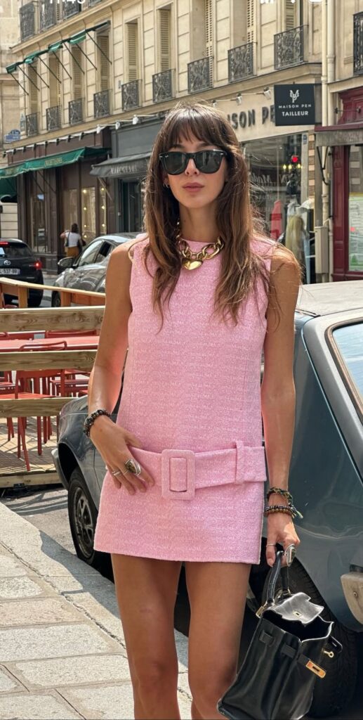 A woman standing on a city street next to a vintage car, wearing a light pink, textured tweed mini shift dress with a wide matching belt and a large rectangular buckle. She is accessorized with dark sunglasses, a chunky gold chain necklace with a heart pendant, and a black leather handbag.