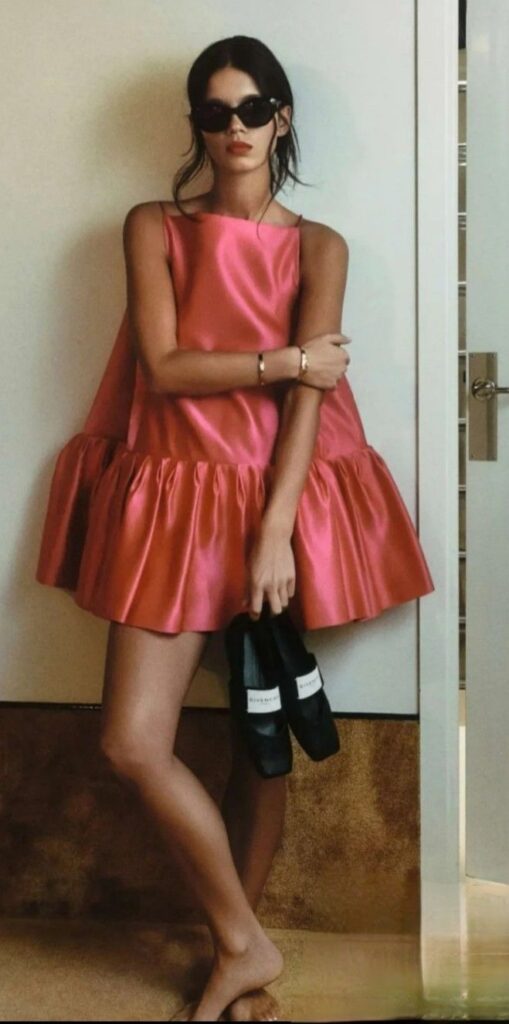A woman posing indoors against a white wall, wearing a voluminous, hot pink satin babydoll mini dress with a straight neckline and a heavy ruffled hem. She is wearing dark sunglasses and holding a pair of black Givenchy slides in her hand while standing barefoot.