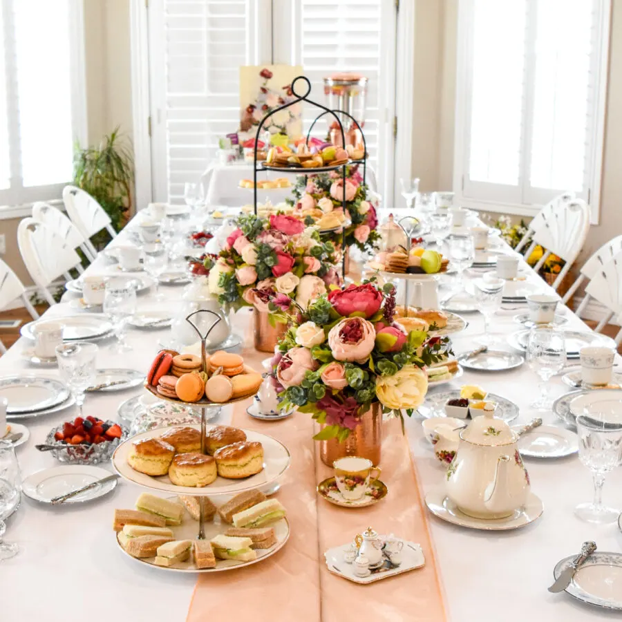 A long table set for a Victorian-style tea party, featuring multiple tiered serving stands with pastries and sandwiches, floral centerpieces, teapots, and fine china place settings on a white tablecloth with a pale pink runner.