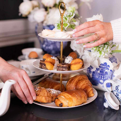 A three-tiered white porcelain stand with a gold handle, filled with breakfast pastries. The bottom tier features flaky croissants and pain au chocolat, the middle tier holds bite-sized brownies and tea cakes, and the top tier has light meringues. White and blue floral china and fresh flowers are visible in the background.