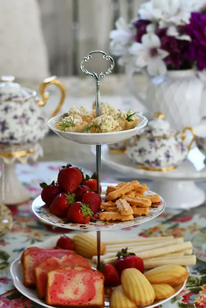 A three-tiered serving stand with a silver handle on a floral tablecloth. The top white tier holds small savory phyllo cups garnished with dill, the middle floral tier features fresh strawberries and cheese straws, and the large bottom tier displays pink marbled cake slices, madeleines, and wafer rolls. Floral teapots and purple flowers are blurred in the background.