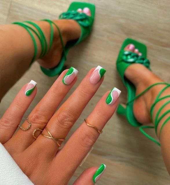 A close-up shot of square-shaped nails featuring a design with green and white wavy lines over a nude base, with the person wearing green strappy heels in the background.