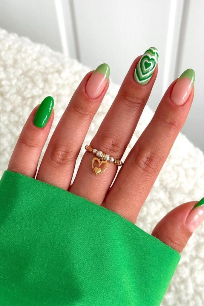 A close-up of a hand with a mix of solid green nails, green French tips, and a ring finger with a layered white and green heart design, wearing a charm ring.