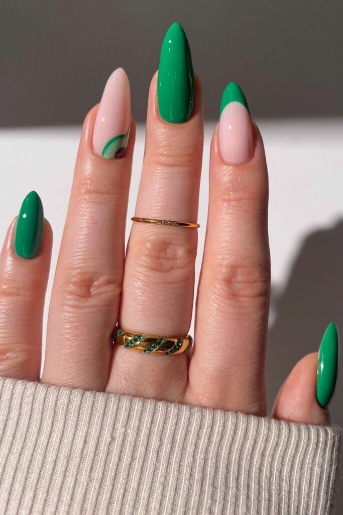 A close-up of a hand with a mix of solid green nails, green French tips, and one accent nail with a small green avocado, wearing gold jewelry.