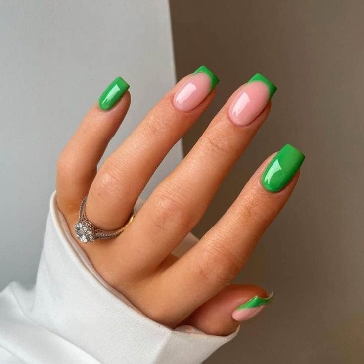 A close-up of a hand with square-shaped nails featuring bright green French tips on a natural pink base, accented by a diamond ring and white sleeve.
