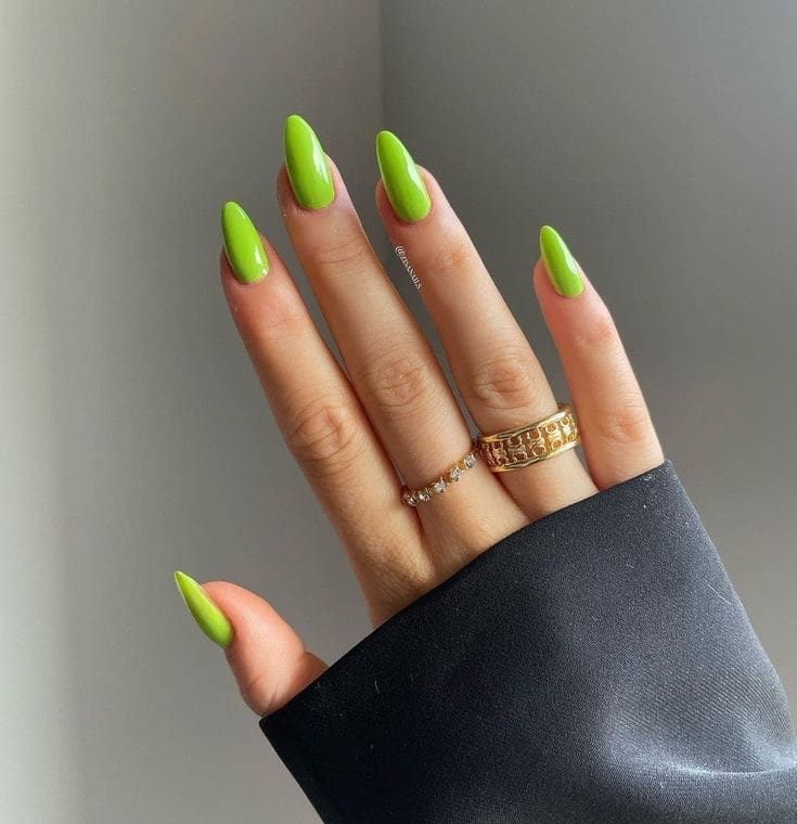 A hand with long, pointed nails painted a solid, bright lime green color, accented by two gold rings and the sleeve of a black jacket.