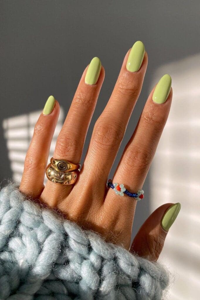 A close-up of a hand with almond-shaped nails painted in a solid, bright lime green color, wearing several gold rings and a blue knitted sweater.