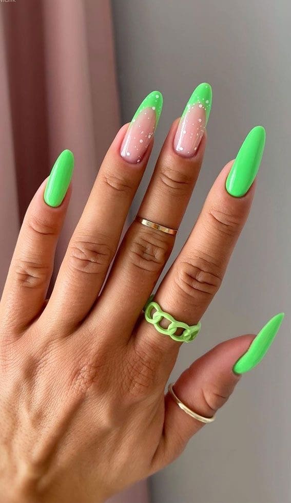 A close-up of hands with almond-shaped nails featuring neon lime green French tips decorated with small white dots, paired with gold rings.