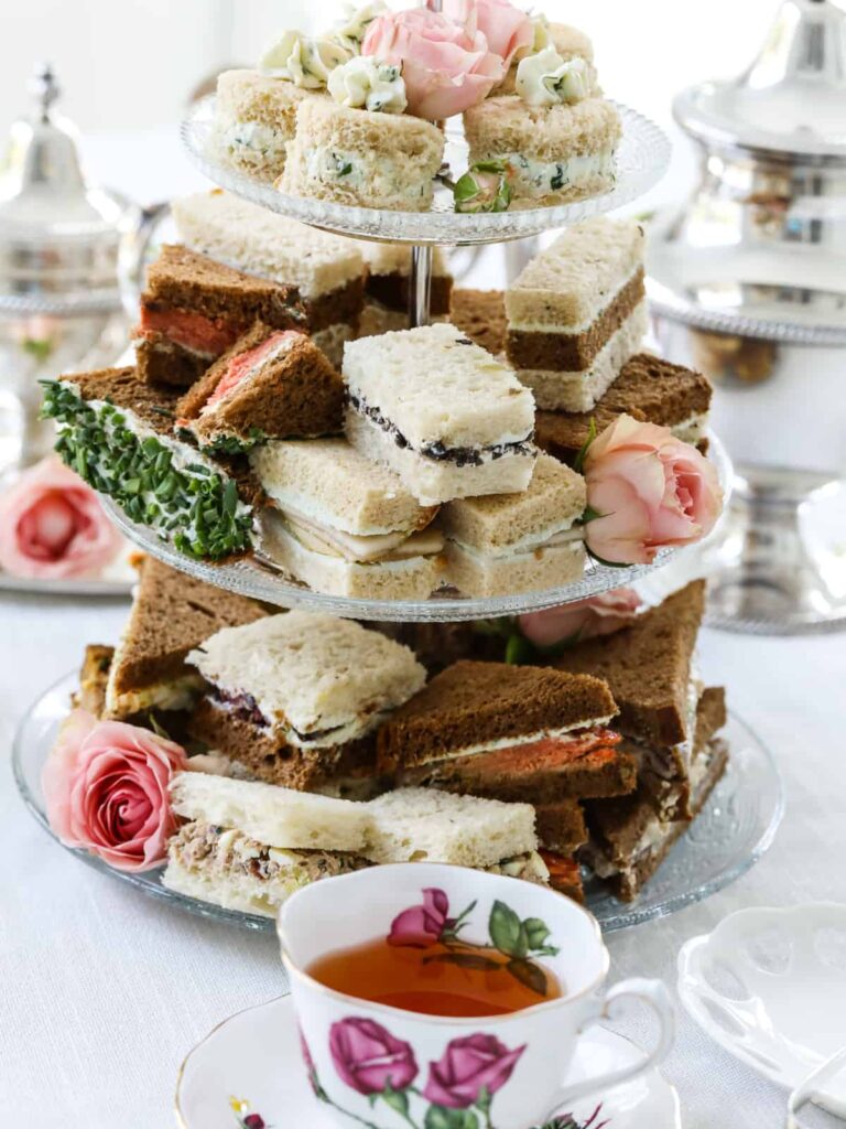 A three-tiered clear glass stand overflowing with assorted tea sandwiches on white and brown bread. Pink roses are tucked between the sandwiches for decoration. In the foreground, a delicate white teacup with pink rose patterns is filled with tea, set against a bright, airy background with silver teapots.