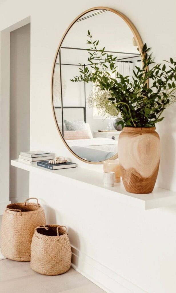 A minimalist white floating entryway shelf underneath a large round gold mirror, featuring an organic wooden vase with green branches and woven baskets underneath.