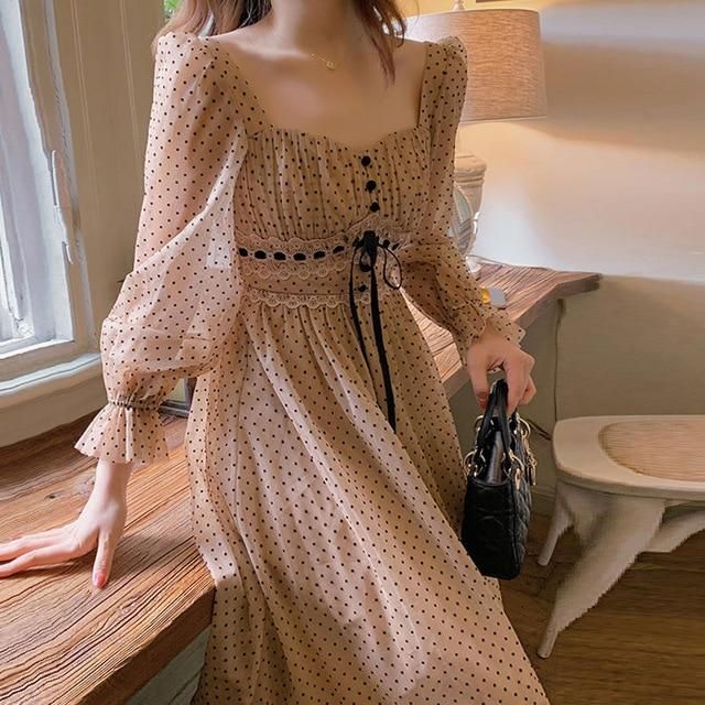 A close-up of a person wearing a modest, long-sleeved beige midi dress covered in a small black polka dot pattern. The dress features a square neckline, a gathered bust with small black buttons, and a defined waistline detailed with black-and-white intricate lace and a dainty black ribbon tie. The person holds a small, structured black quilted handbag against a warm, indoor setting with a wooden table.
