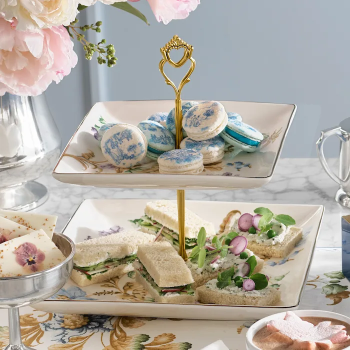 A two-tiered square ceramic serving stand with a gold heart-shaped handle. The top tier features light blue macarons with a vintage toile pattern, and the bottom tier holds assorted finger sandwiches garnished with radishes and sprouts. The stand sits on a marble surface next to fresh pink peonies and silver serveware.