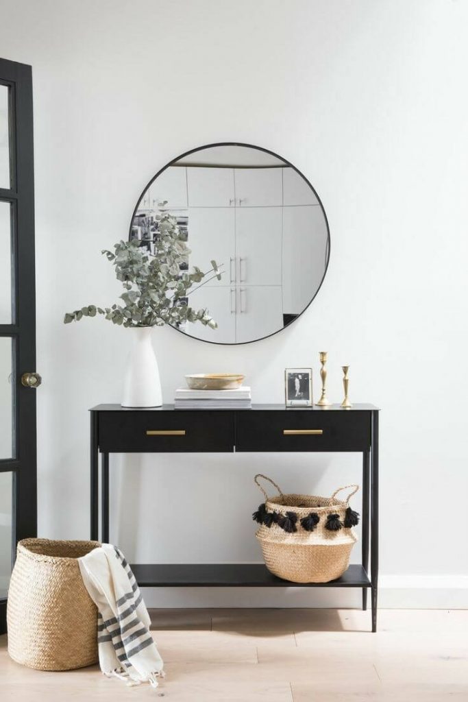 A contemporary black entryway console table with gold handles, a large round black mirror, a white vase with eucalyptus, and a woven basket underneath.