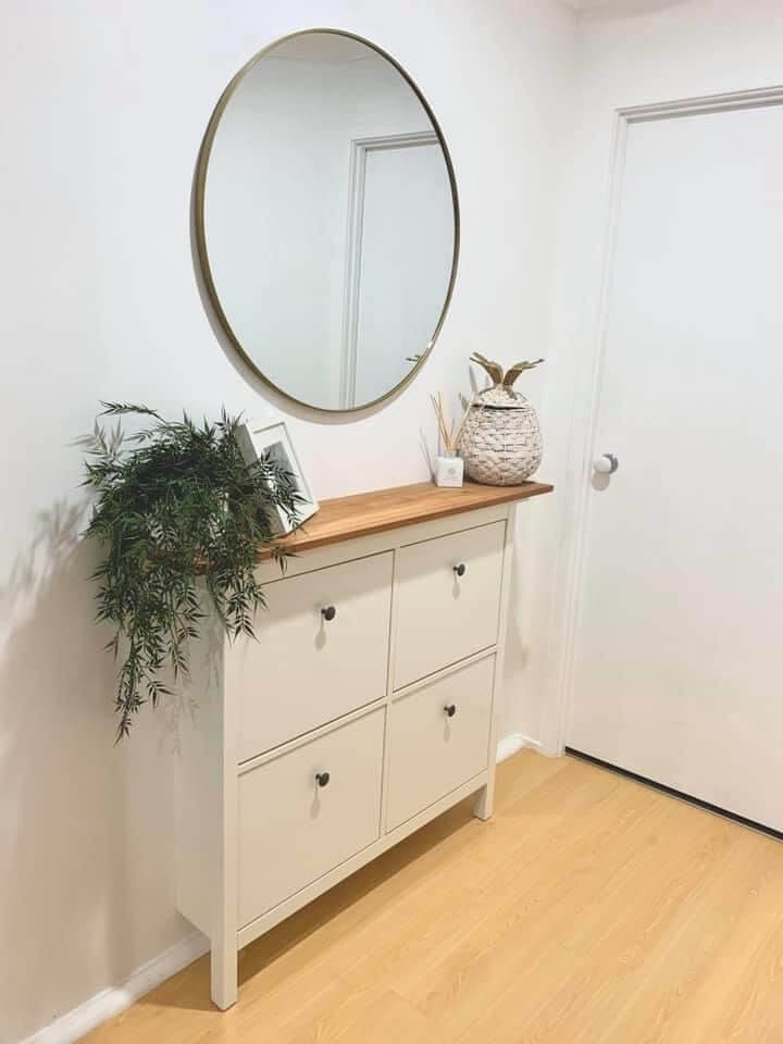 A narrow white Scandinavian-style shoe storage cabinet in a hallway with a wooden top, trailing green plant, and a large round brass mirror hanging above.