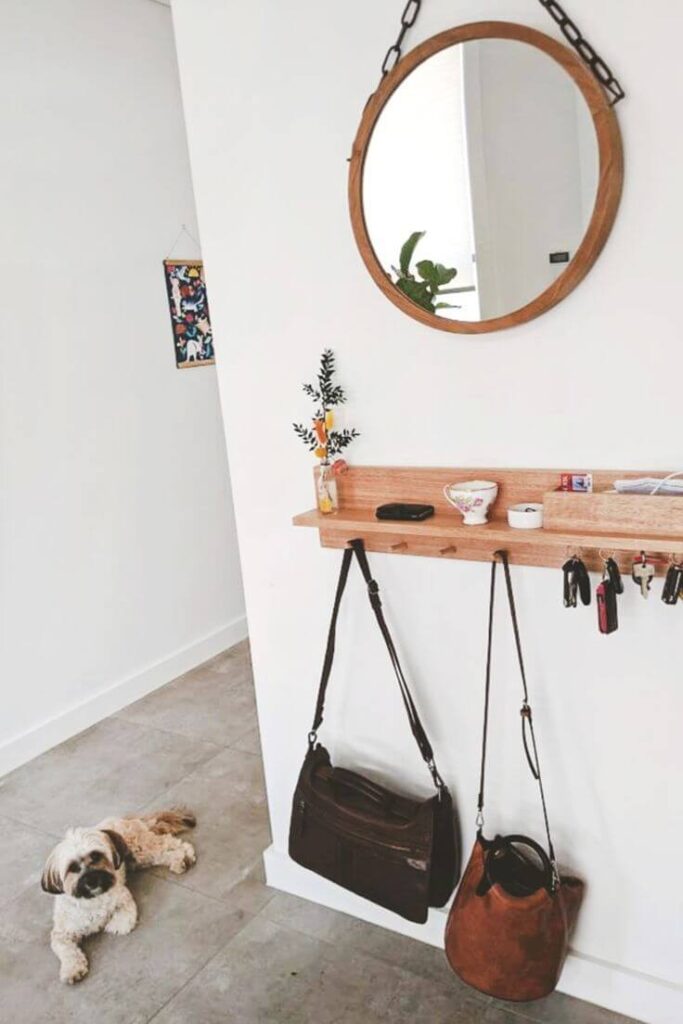 A small wall-mounted light wood entryway shelf with built-in hooks for hanging keys and leather bags, topped with a small vase and decorative teacup under a round wooden mirror.