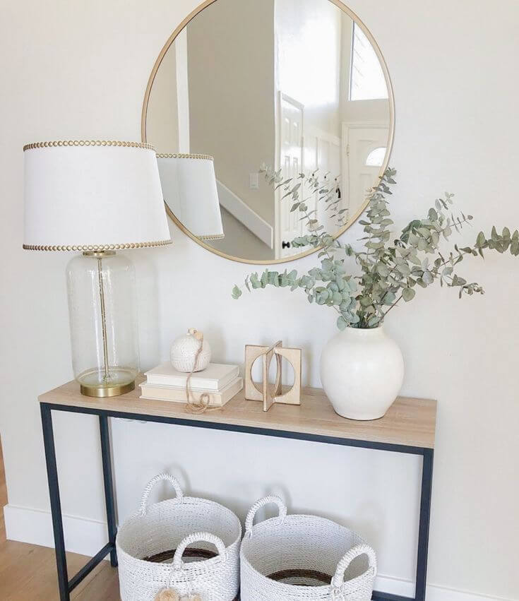A bright, neutral styled entryway table with a glass table lamp, a round gold mirror, a white ceramic vase filled with eucalyptus, and two white woven storage baskets below.