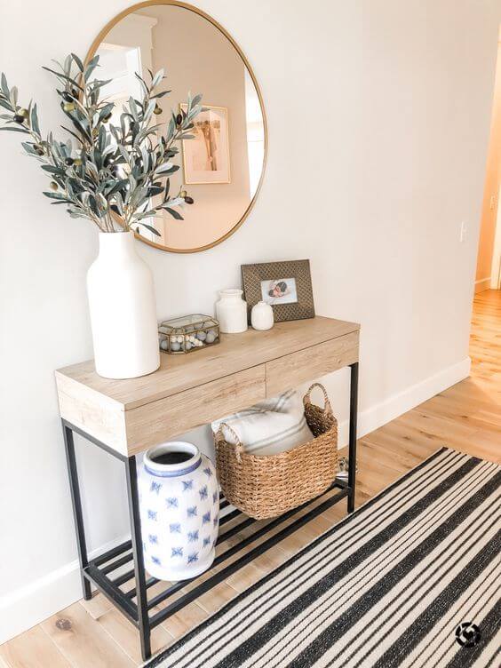 A light oak and black metal console table decorated with an olive branch arrangement in a white vase, stacked books, and a woven basket on the bottom shelf.