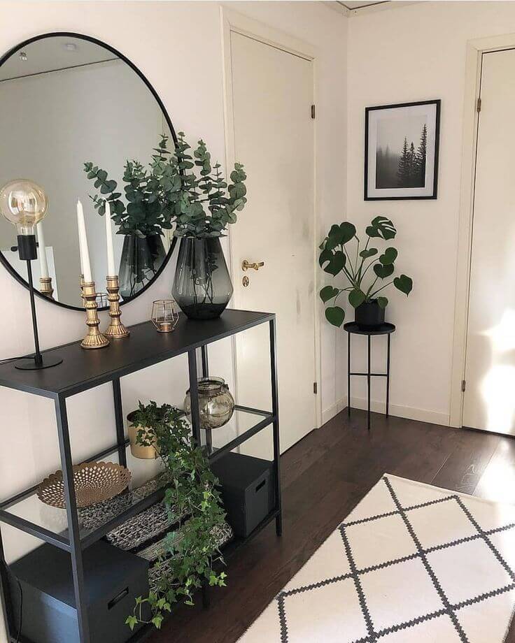 A tiered black metal entryway console table heavily styled with eucalyptus in a smoked glass vase, brass candlesticks, trailing ivy, and a large round black mirror.