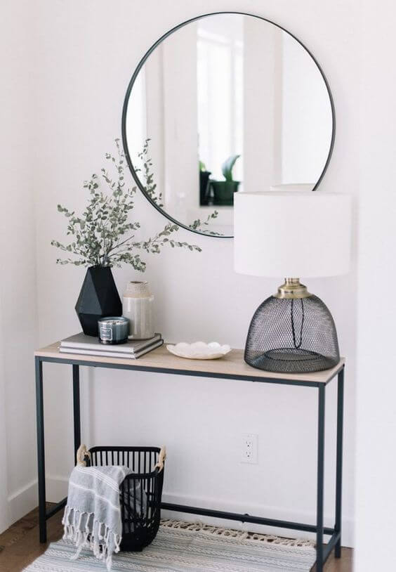 A modern industrial entryway table with a black metal frame, a large round black-trimmed mirror, a geometric vase with eucalyptus, and a black mesh table lamp.