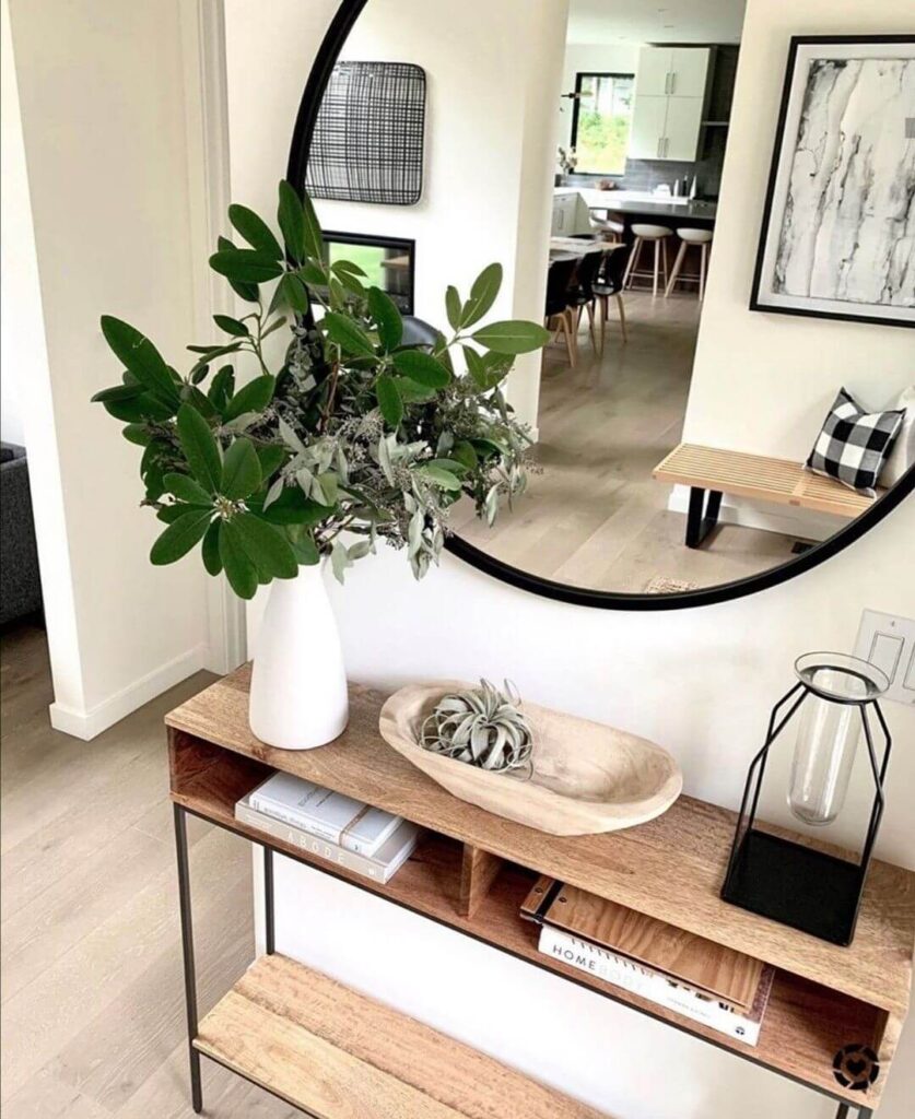 A wooden entryway console table with built-in shelves featuring stacked books, a white vase with large green branches, a wooden dough bowl, and a large round black mirror above.