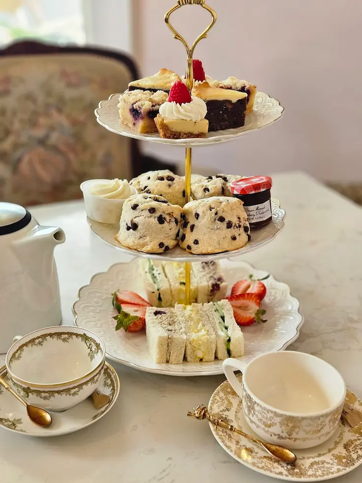 three-tier stand holds tea sandwiches and strawberries on the bottom, scones and jam on the middle, and various mini desserts and raspberries on the top. Teacups and a teapot surround it on a marble table.