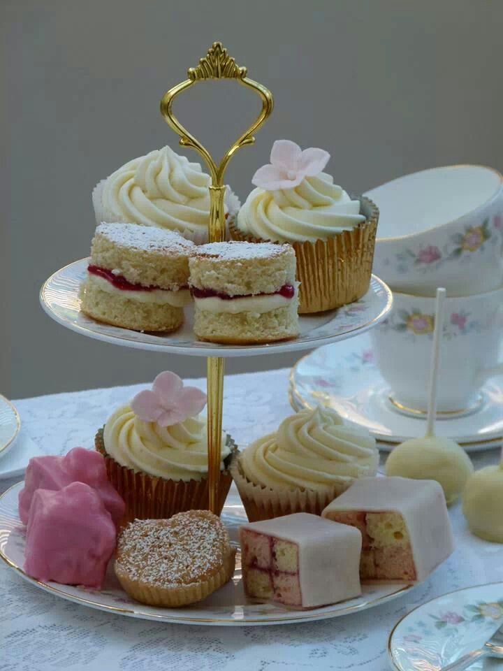 A two-tier ceramic stand with a gold handle displays elegant mini cakes, cupcakes, and macarons. It sits on a white lace tablecloth with stacked teacups in the soft-focus background.