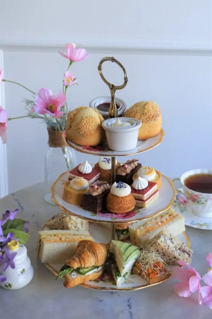 A three-tier ornate stand displays small finger sandwiches on the lower level, a selection of intricate, colorful pastries and tarts on the middle level, and two large scones on the top, with tea and pink flowers adjacent.