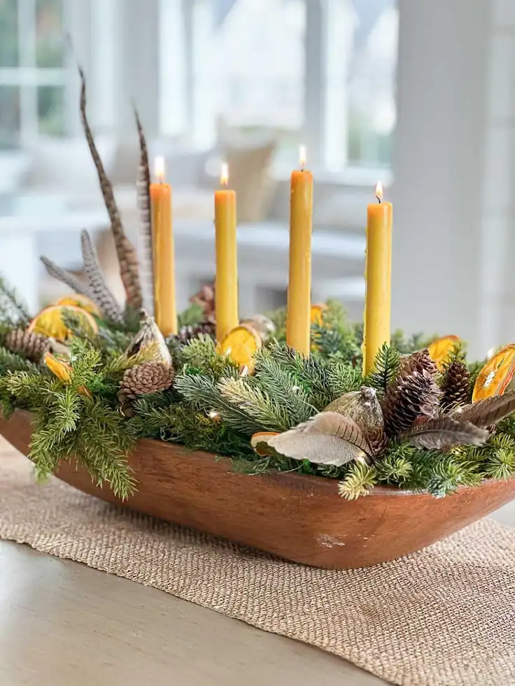 Wooden dough bowl centerpiece with lit mustard taper candles, fir greenery, dried orange slices, pheasant feathers, and pinecones.