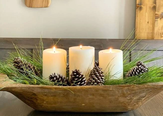 Three lit ivory pillar candles surrounded by long pine sprigs and natural pinecones inside a wooden dough bowl centerpiece.