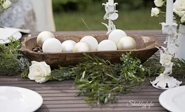 Wooden dough bowl filled with oversized cream and gold ornament balls, surrounded by rosemary, juniper, and white roses on an outdoor table.