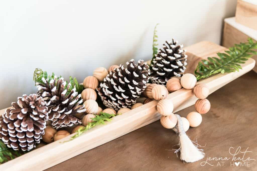 Long wooden dough bowl filled with a wood bead garland, snow-tipped pinecones, and faux fern sprigs for a winter table runner.