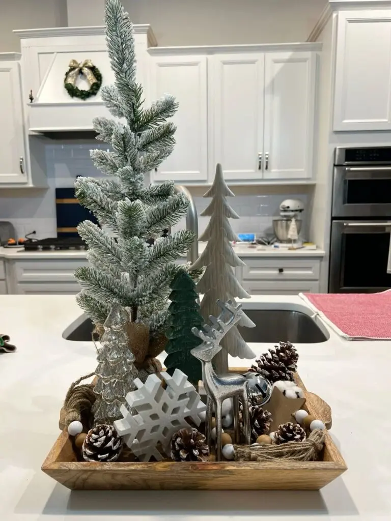 Wooden dough bowl on a kitchen counter with flocked mini Christmas trees, pinecones, wood beads, cotton stems, and a silver reindeer figurine.