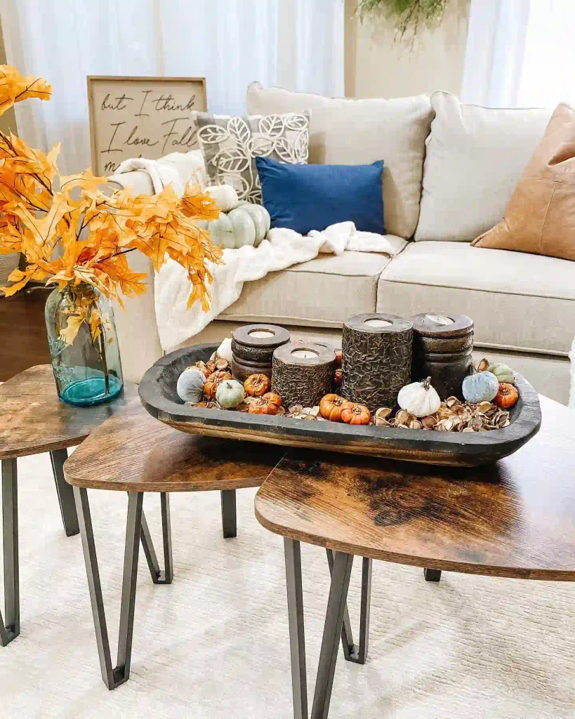 Black farmhouse dough bowl on a wood coffee table filled with carved wood candle holders, mini velvet pumpkins, and dried botanicals beside a vase of orange fall leaves