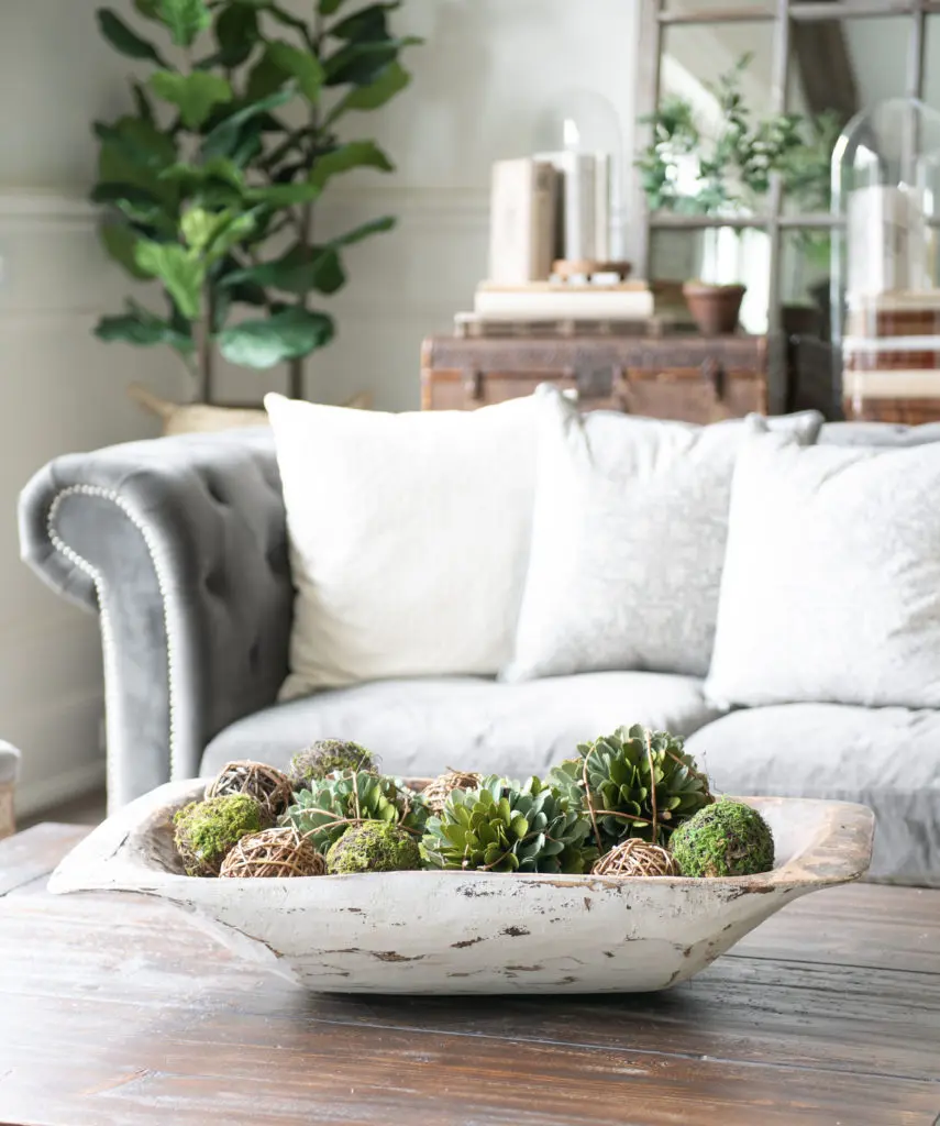 Whitewashed farmhouse dough bowl on a rustic wood coffee table filled with moss balls, grapevine spheres, and faux succulents in front of a gray tufted sofa