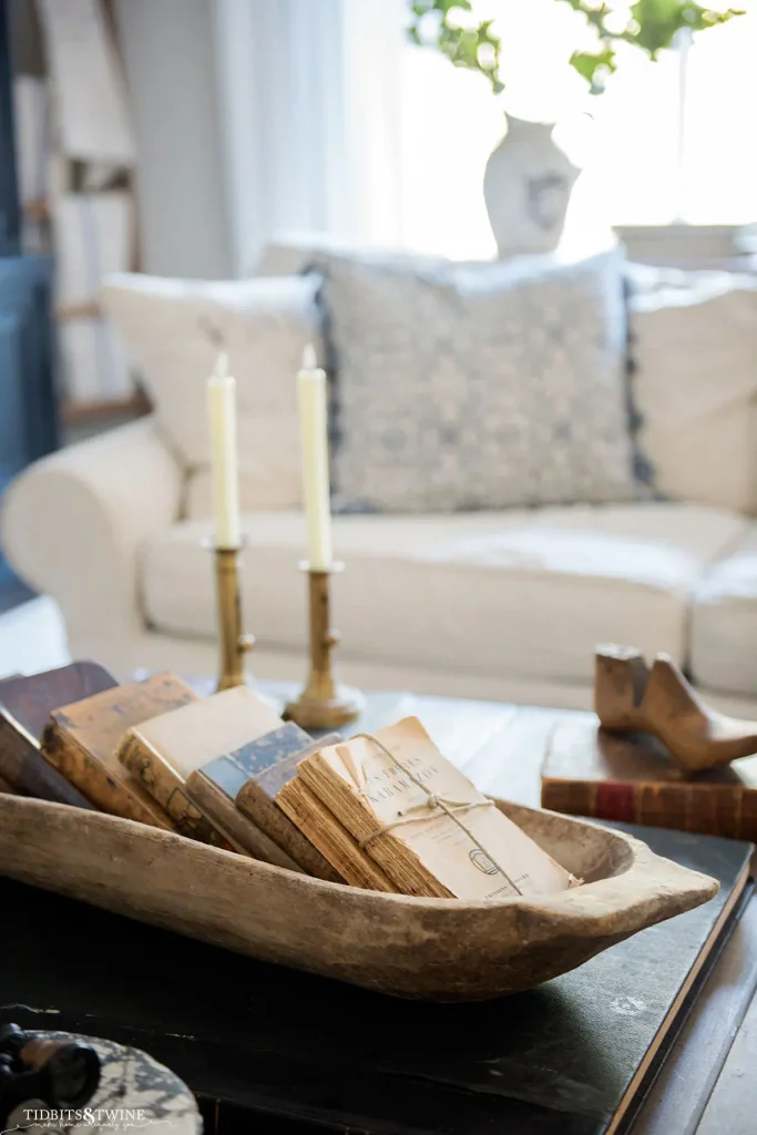 Weathered wood dough bowl filled with vintage books tied with twine on a dark wood coffee table with brass candlesticks and cream taper candles in front of a cream sofa
