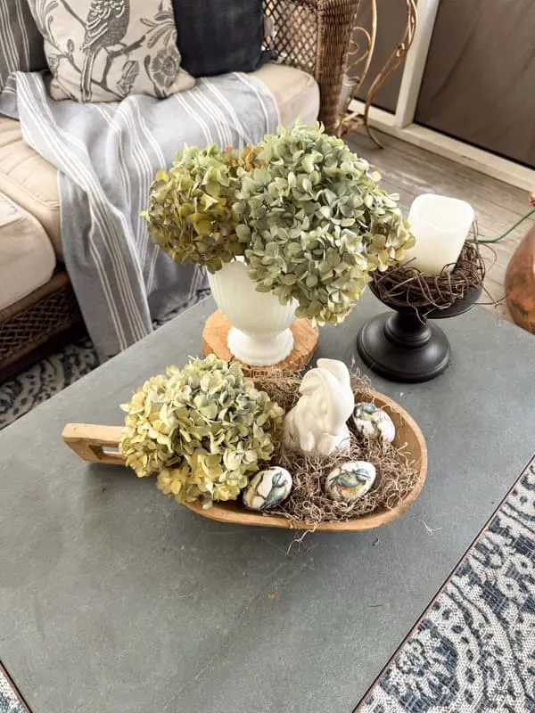 Wood dough bowl with handle on a coffee table filled with moss, dried green hydrangeas, a white ceramic bunny, and bird-print eggs beside a milk glass vase of dried hydrangeas and a candle in a twig nest