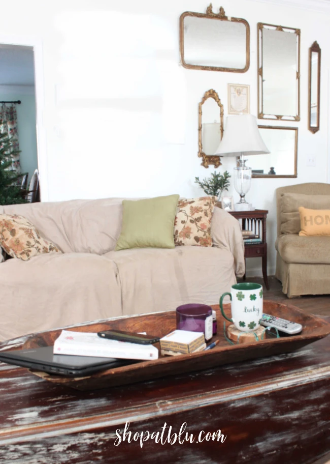 Wood dough bowl on a distressed coffee table used as a tray holding a laptop, phone, book, notepad, candle, remote, and mug on a wood slice coaster in front of a neutral sofa